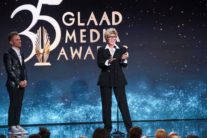 Person on stage at GLAAD Media Awards speaking, another person on left looking on, audience in foreground