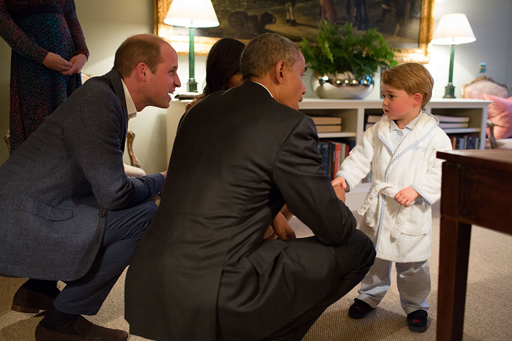 Prince William kneels beside President Obama who is chatting with Prince George in a white robe, in an elegant room