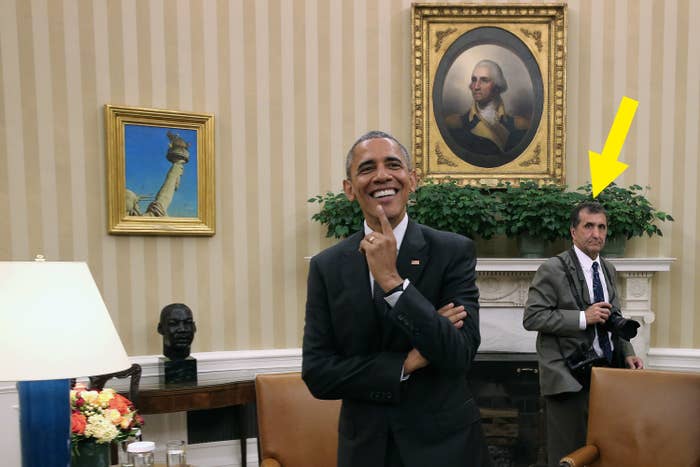 President Obama in a suit smiling with hand on chin, with Pete behind him with camera, in the Oval Office