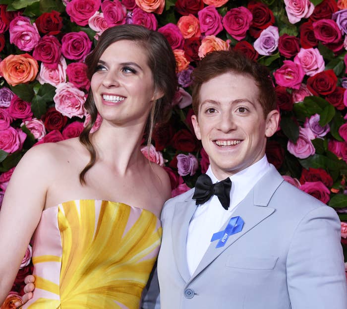 Two people standing in front of a floral backdrop, one in a strapless dress and the other in a light suit with a bow tie