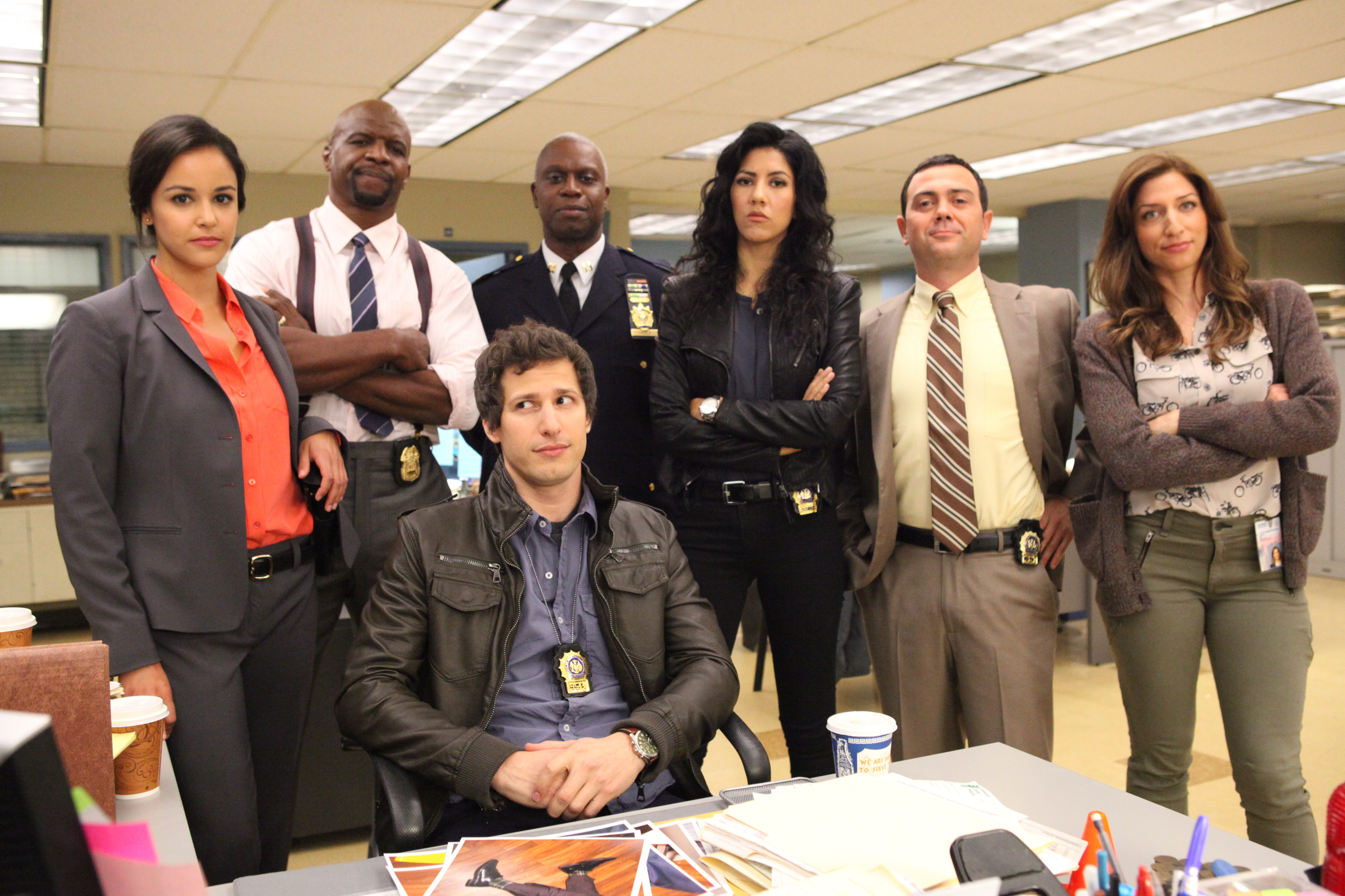 Cast of Brooklyn Nine-Nine posing at a precinct desk: Amy in a suit, Terry in a dress shirt, Holt in a suit, Rosa in leather jacket, Boyle in a tie, and Diaz in a jacket