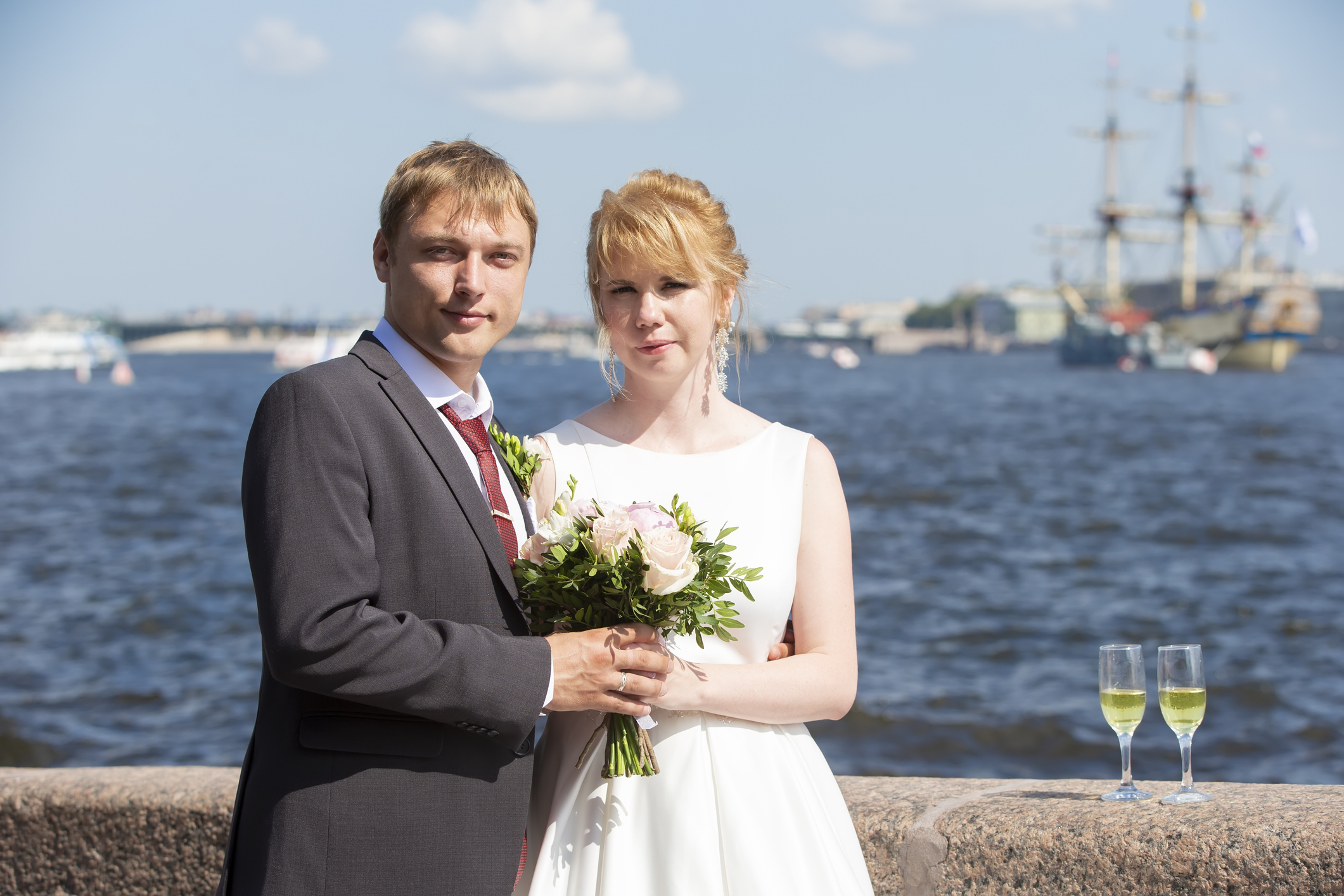 Bride and groom holding hands by the river with a ship in the background, bride holding a bouquet, and two glasses on the ledge