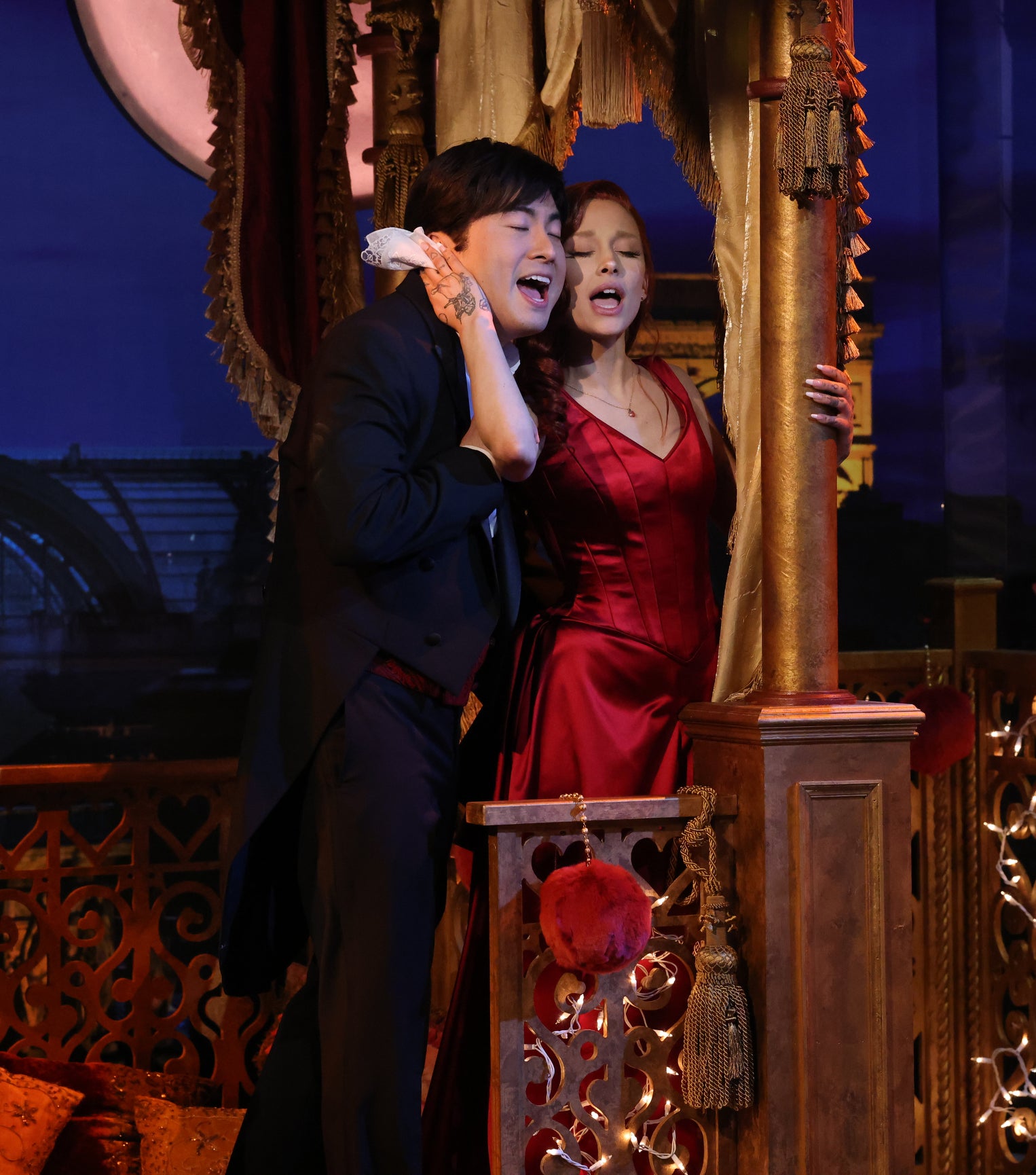 Two performers on stage, one in a suit and the other in a red dress, sharing a moment in a musical production