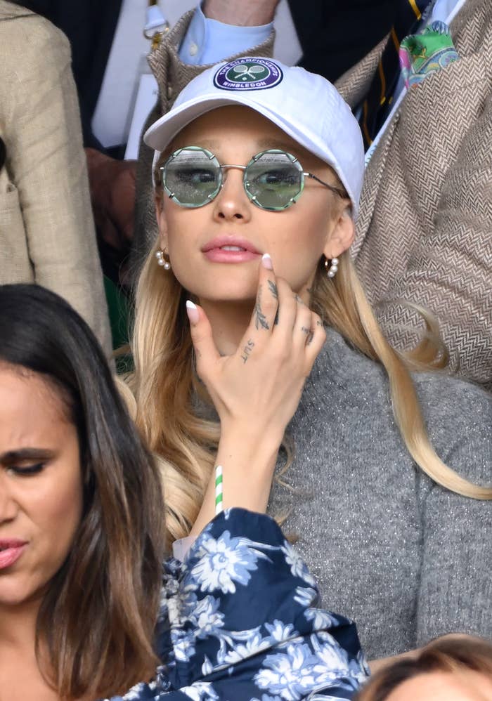 Woman with cap and sunglasses touching her face, surrounded by crowd