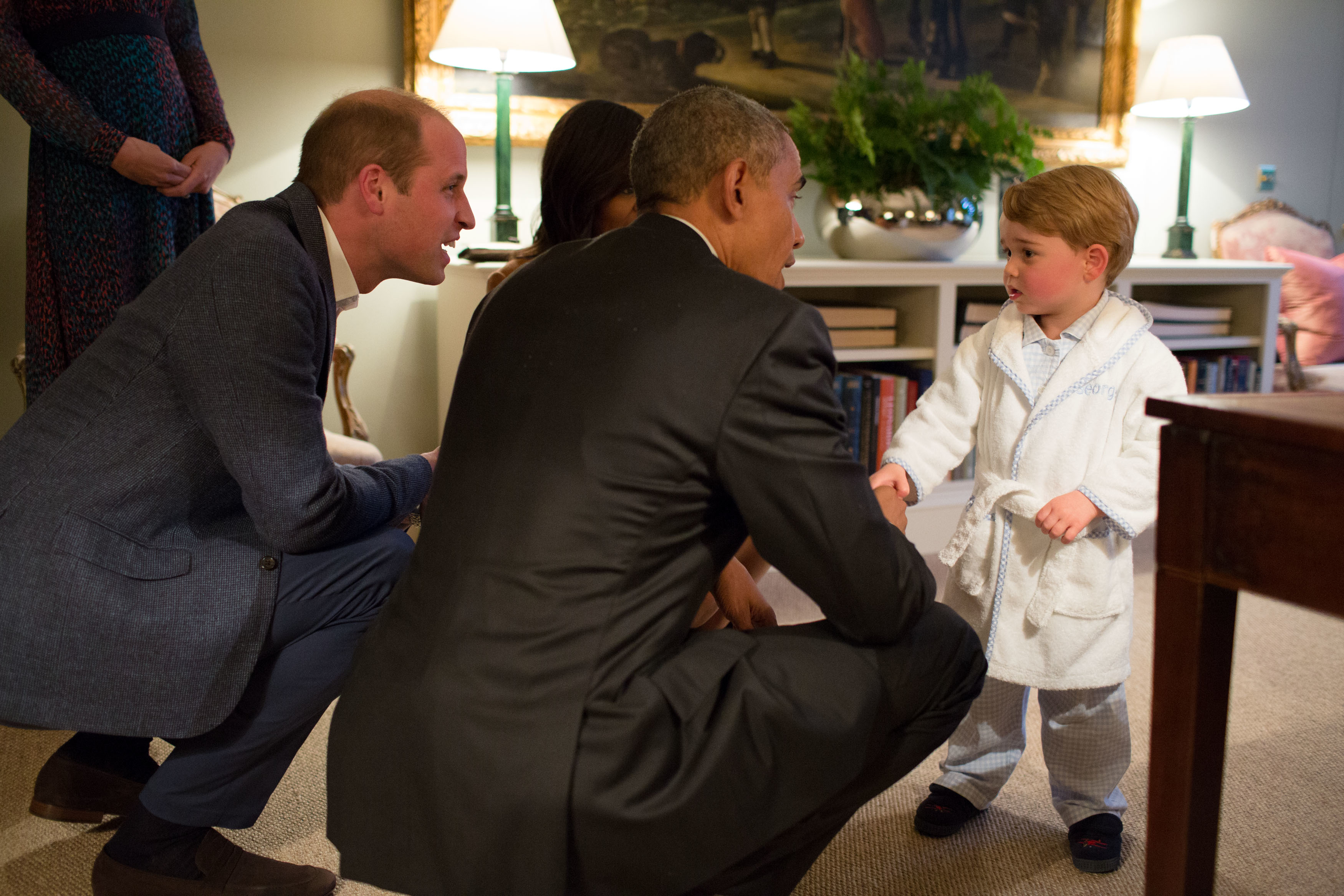 Prince William, President Obama, and Prince George in a room; George wears a white robe; the adults are smiling down at him