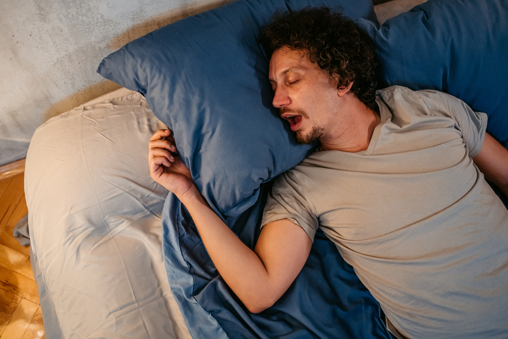 Man sleeping peacefully with one arm extended, lying on a bed with pillows