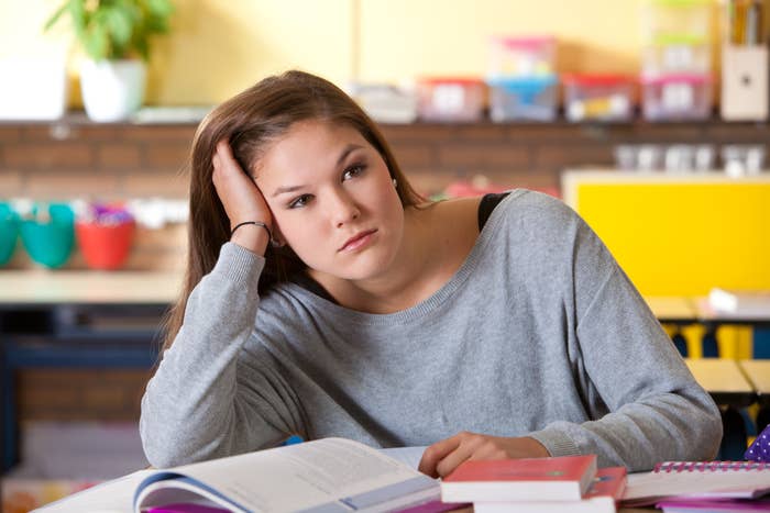 A student rests her head on her hand, looking away from a book on a desk in a classroom