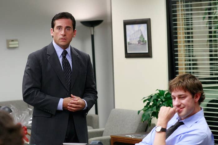 Two men in an office, one standing in a suit, the other sitting and smirking