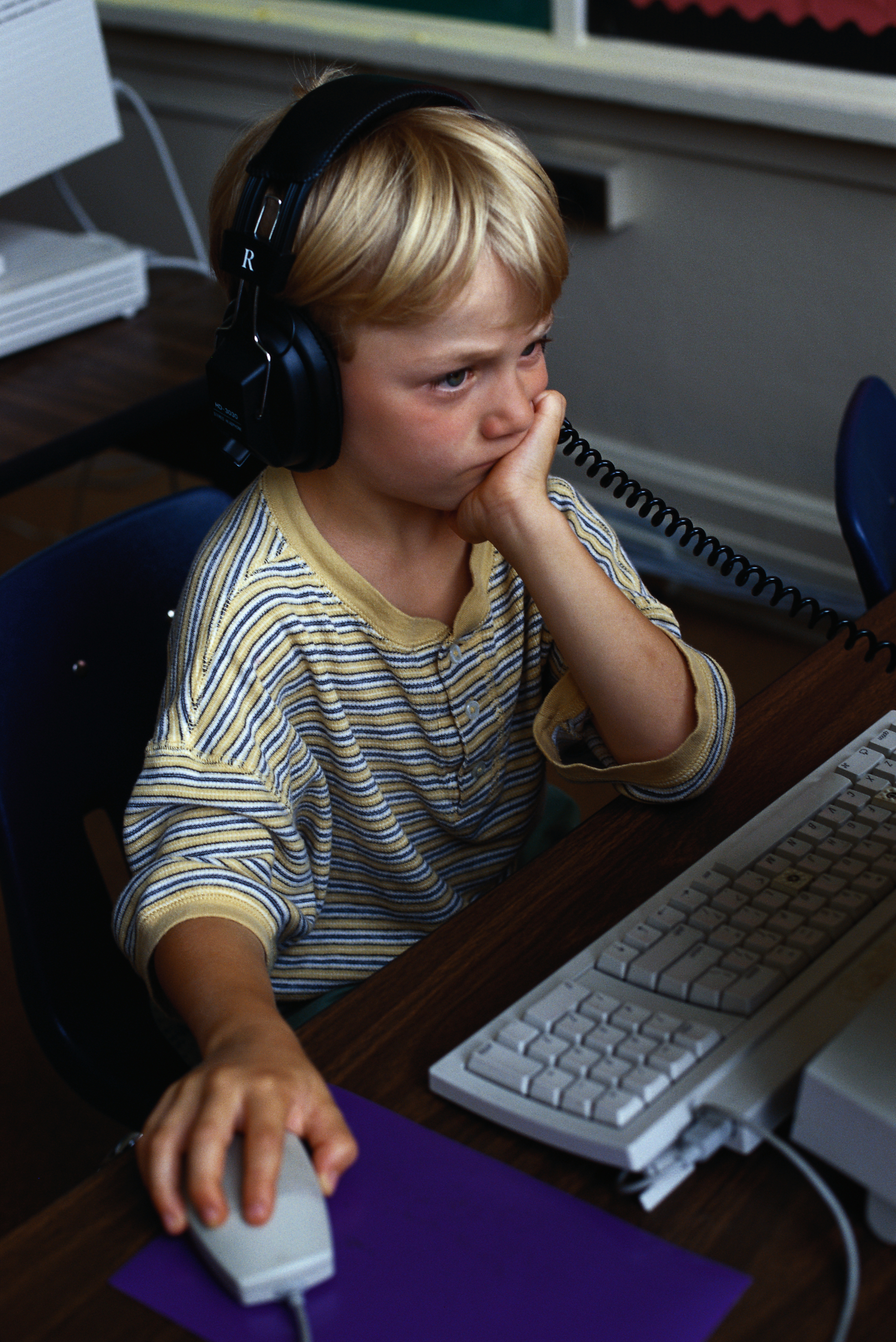 Child in a striped top uses a computer mouse and wears headphones, looking focused