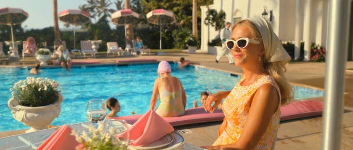 Woman in sunglasses by a pool with diners and swimmers, wearing a patterned dress, seated at a table with a pink napkin