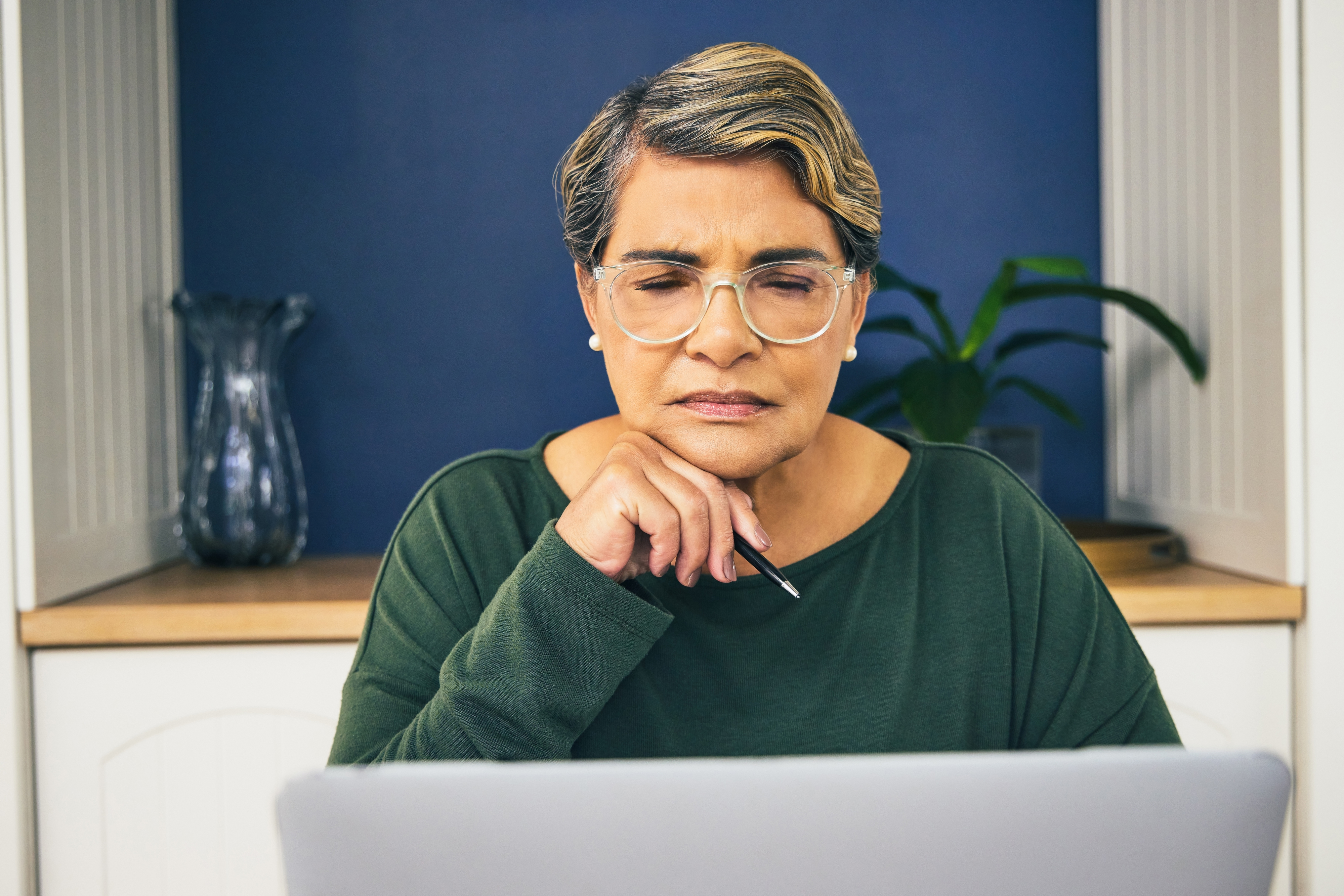Person at a laptop appears contemplative or focused, resting chin on hand, wearing glasses and a top