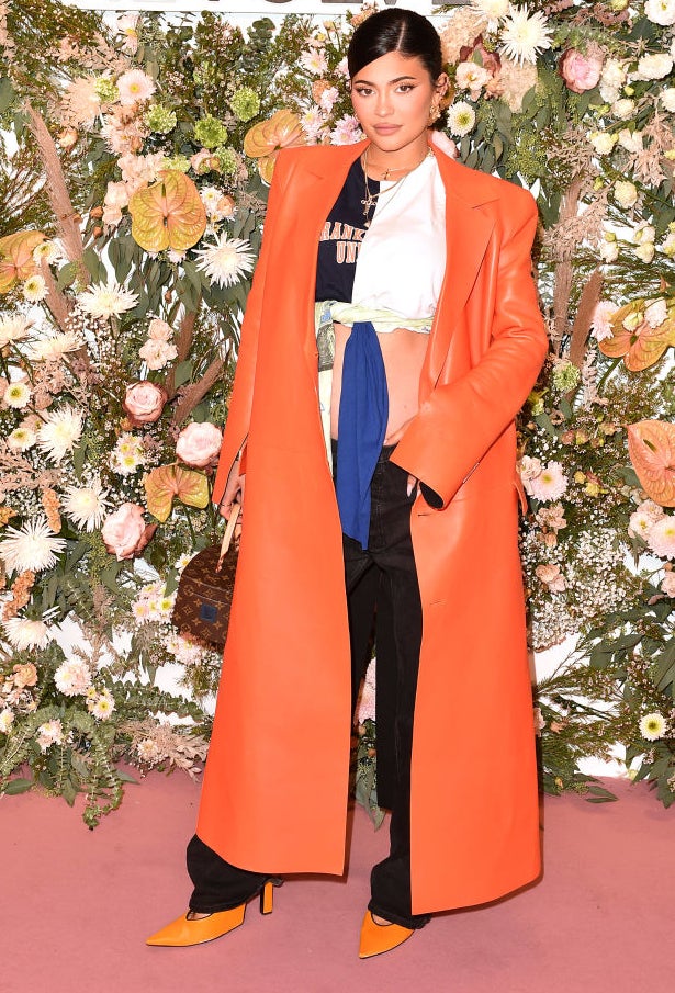 Kylie at event in bright coat over graphic tee and pants, standing before a floral backdrop