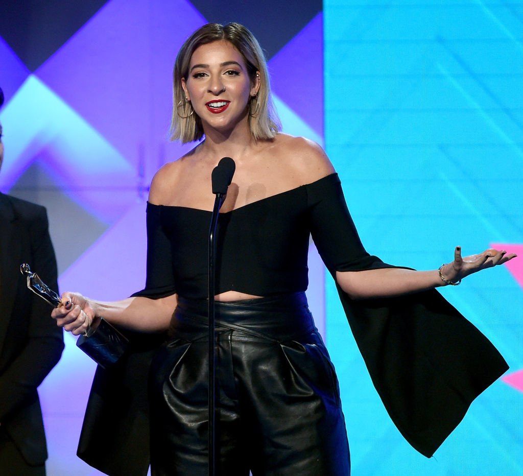 Gabbie on stage with award, wearing off-the-shoulder top and leather pants, speaking into a microphone