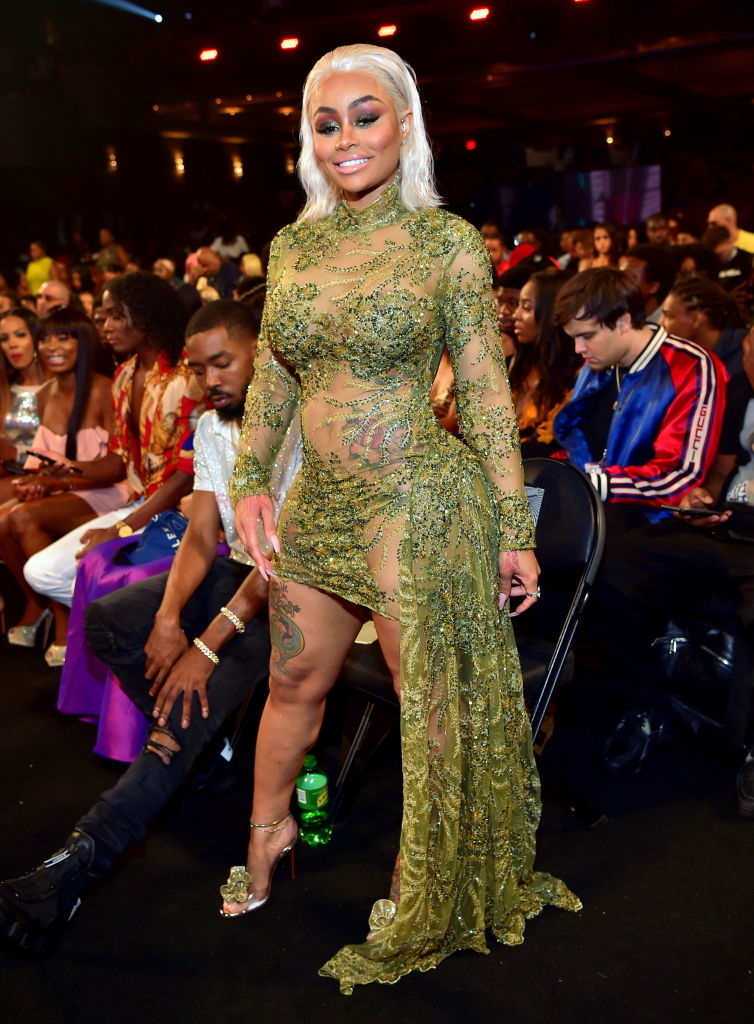 Blac Chyna in a sheer, embellished long-sleeve gown at an event, posing with hand on hip