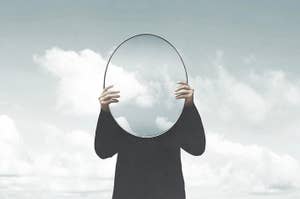 Person holding a mirror reflecting clouds, head obscured by the mirror's reflection
