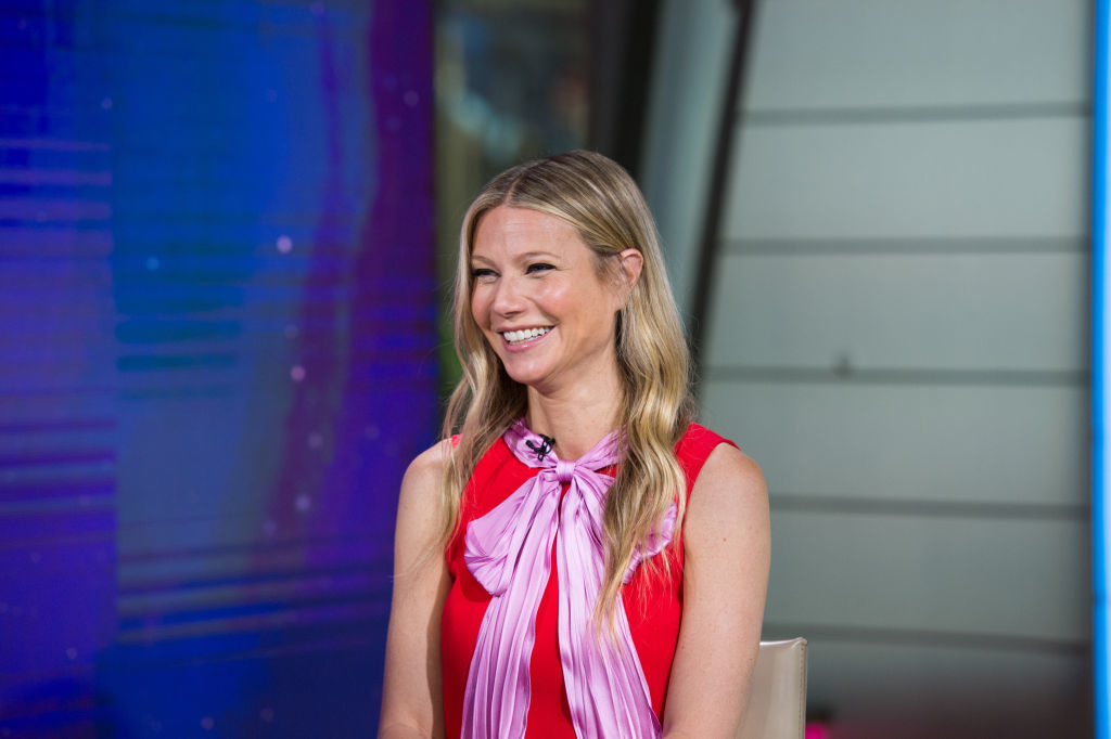 Gwyneth on a talk show smiling, wearing a sleeveless top with a necktie detail