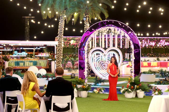 Woman in red dress stands at wedding altar on reality show set, audience seated facing her
