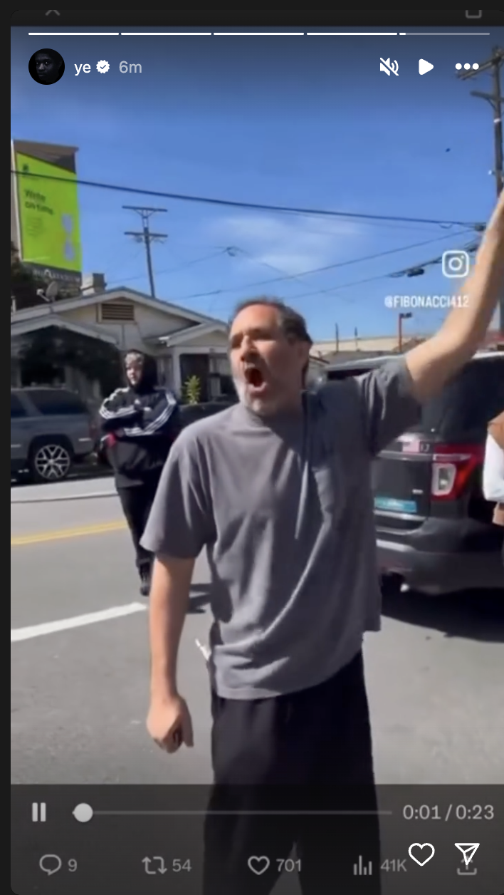 Person in gray attire yelling on a street with cars and bystanders behind