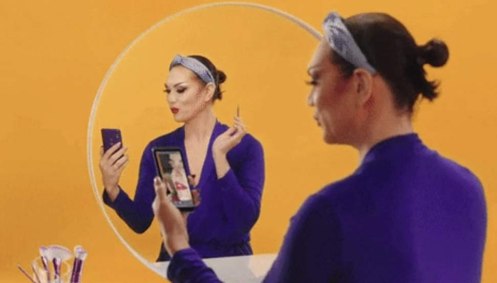 Woman applies makeup while looking at her phone, with reflection in mirror