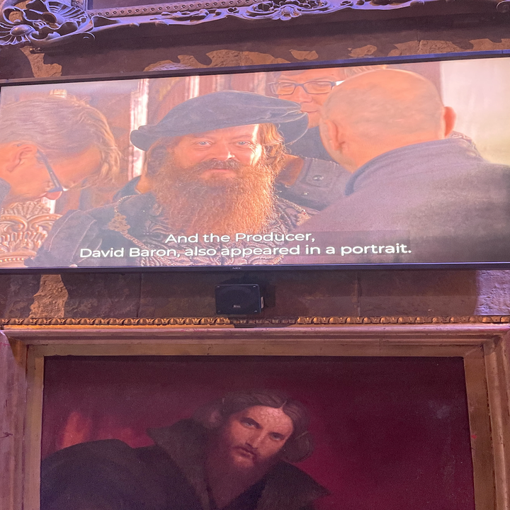 TV screen showing a bearded man in a beret captioned with David Baron's cameo, portrait painting below