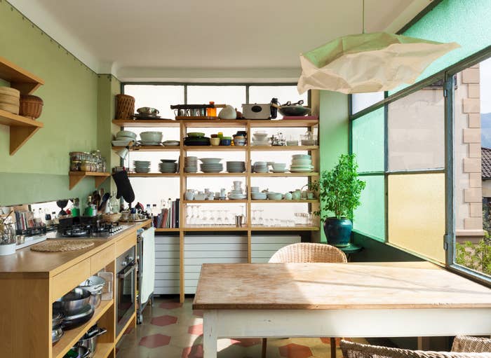 A tidy kitchen with open shelves, various utensils, a central table, and greenery  near the window