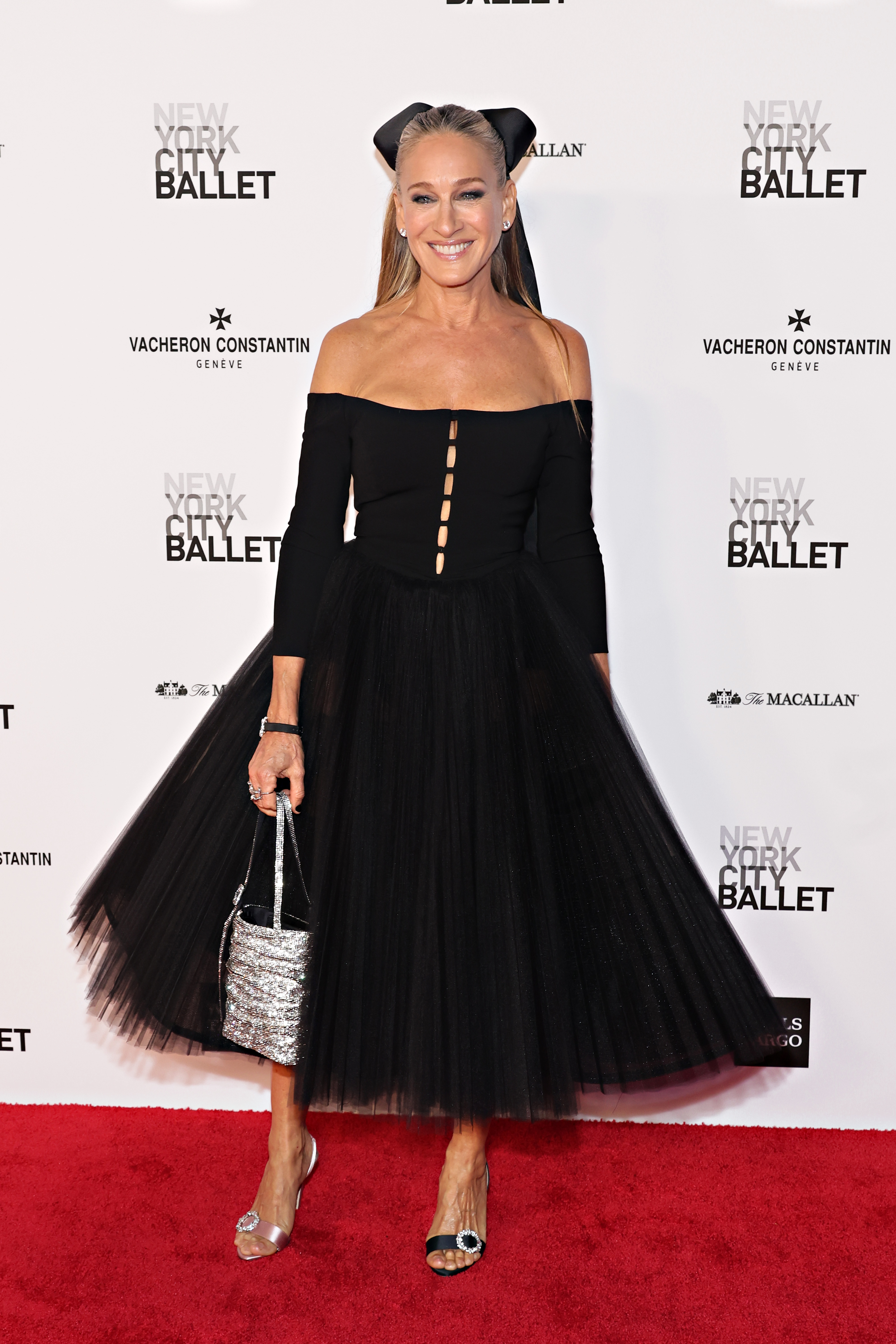 Sarah Jessica Parker at an event wearing a black off-the-shoulder dress with tulle skirt and carrying a glittery handbag