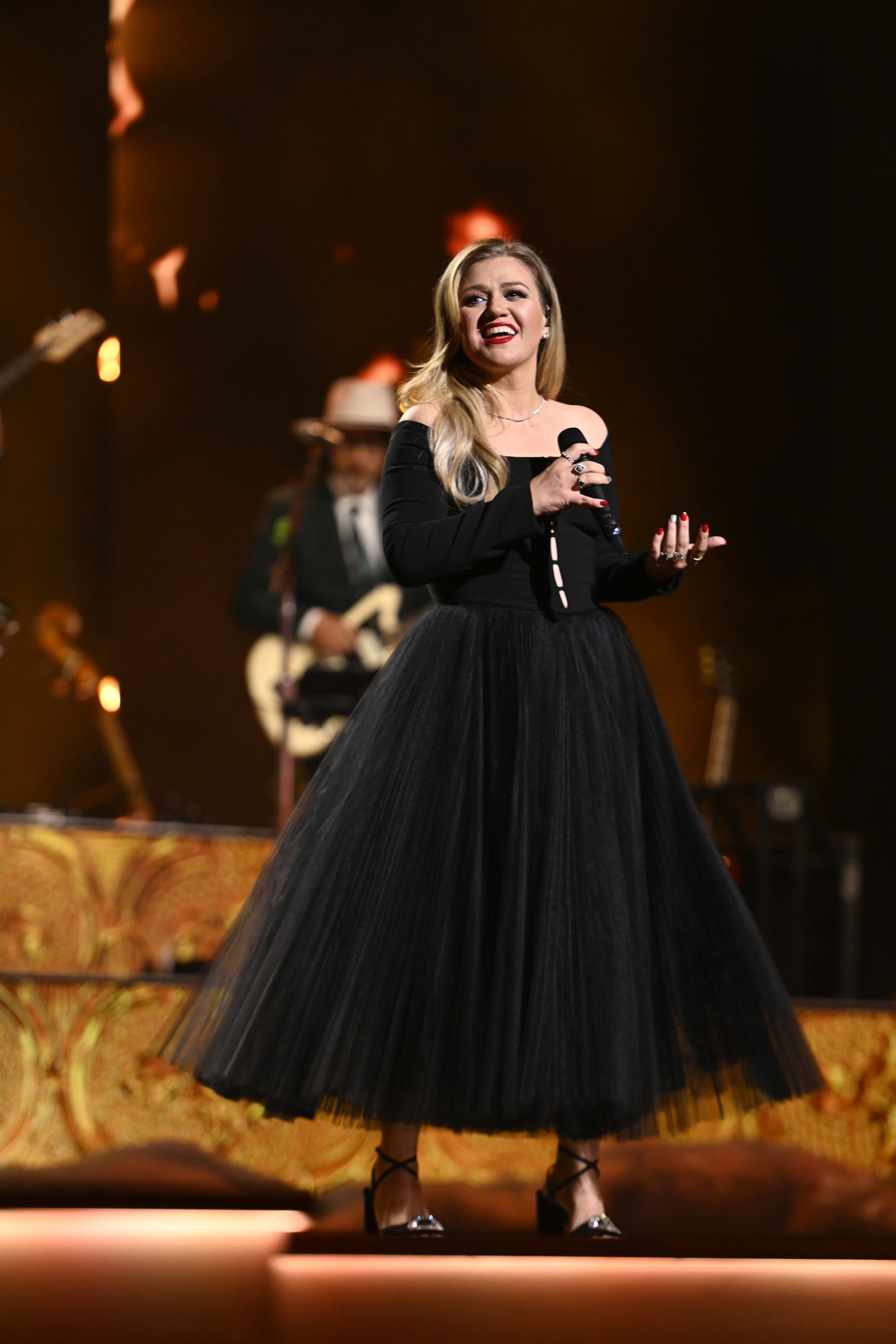 Kelly Clarkson performing on stage in a black tulle dress and heels; a musician is in the background
