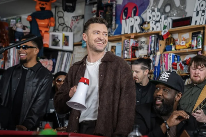 Justin Timberlake with a megaphone, flanked by a band in a room with eclectic decor