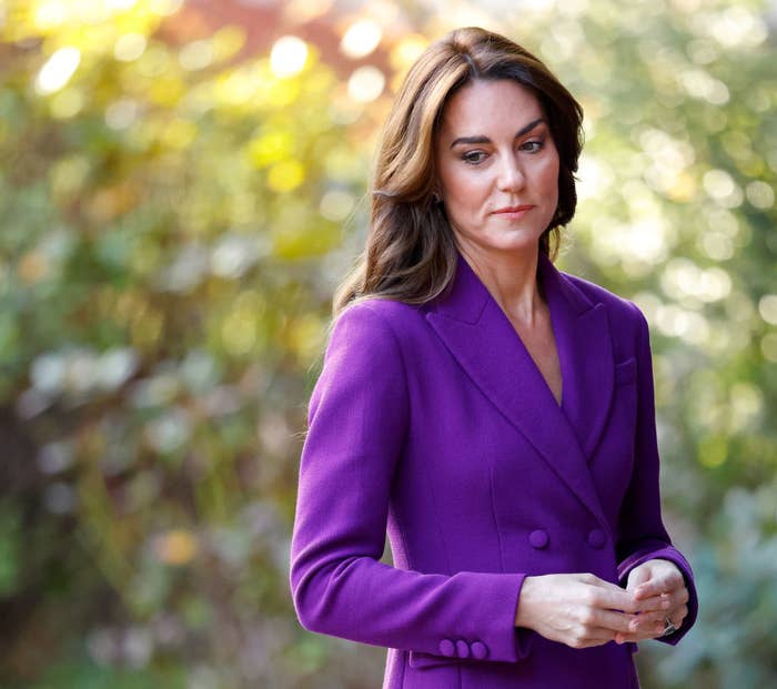 Kate in a formal blazer stands with a thoughtful expression, hands clasped in front