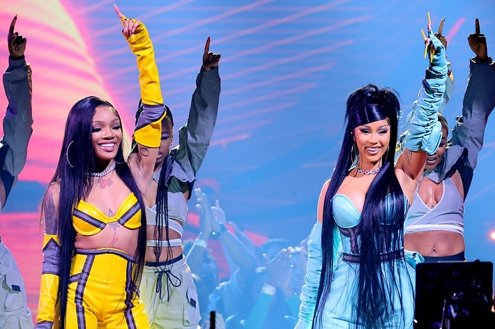 Two performers on stage with raised hands, one in a yellow outfit, the other in blue, with backup dancers