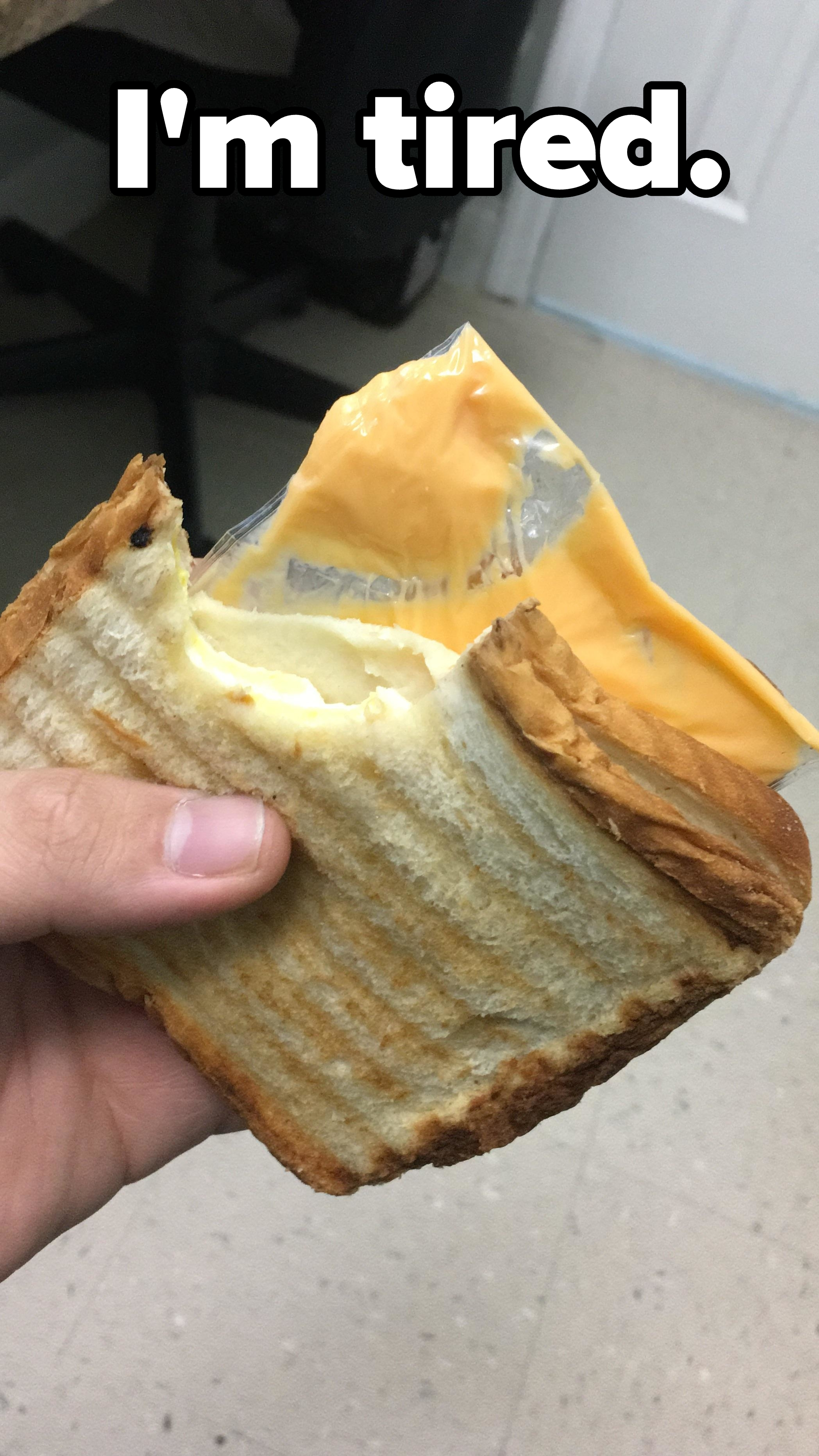 Hand holding a grilled cheese sandwich showing a slice of American cheese still in the plastic, with a bite taken out