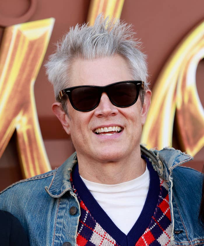 A closeup of him in a denim jacket and sunglasses smiling at a daytime event
