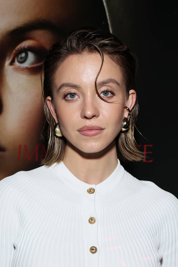 closeup of her with a slicked back hairstyle, wearing a ribbed top with gold buttons, and gold hoop earrings