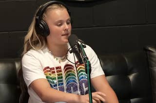 JoJo Siwa in a radio interview, speaking into a microphone, with screens in the background