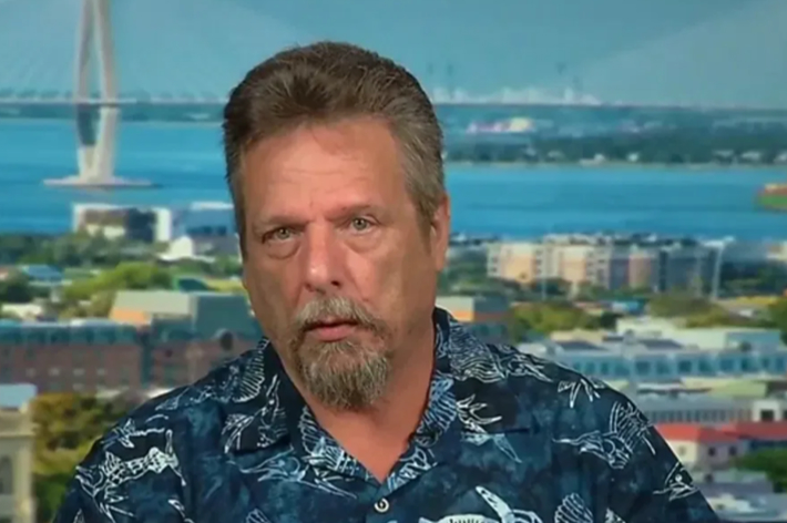 John McAfee wearing a blue-patterned shirt during an interview with a bridge and water in the background