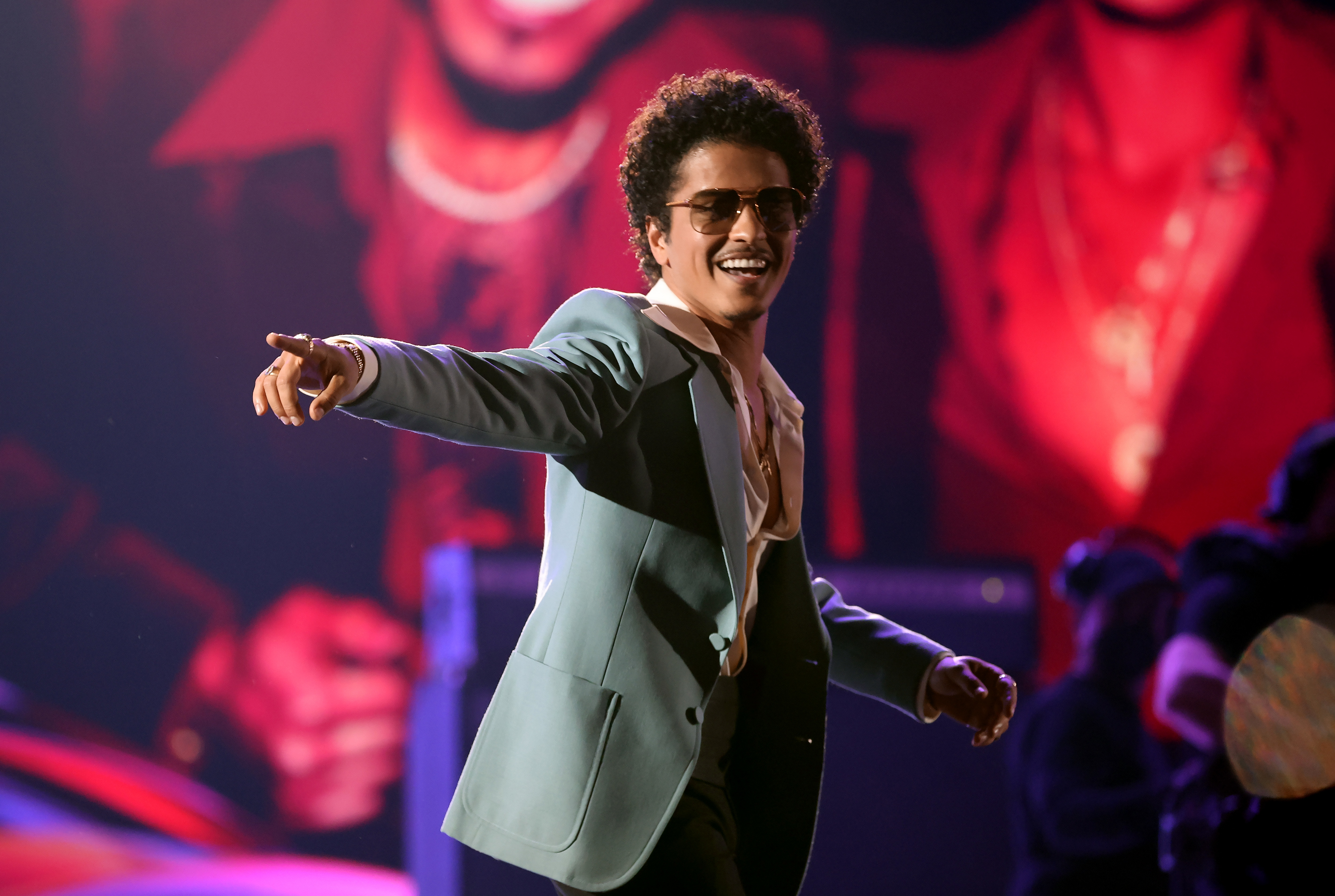 Bruno Mars performing on stage, wearing a stylish suit and sunglasses, gesturing to the audience