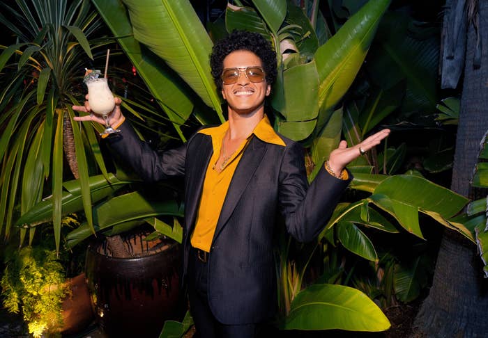 Bruno Mars posing with a drink, sporting a black blazer over a yellow shirt. He&#x27;s smiling against a backdrop of tropical plants