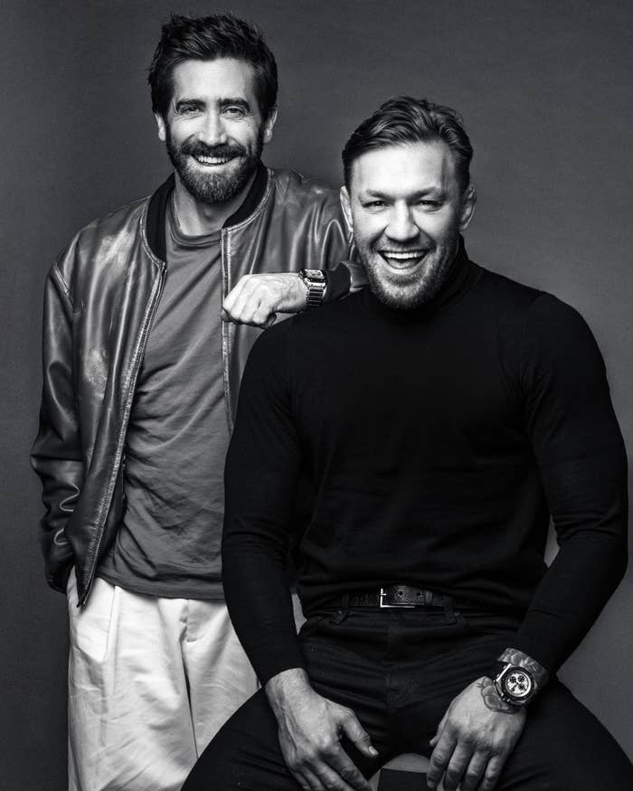Two men smiling, one seated in a turtleneck and slacks, the other standing in a jacket and jeans