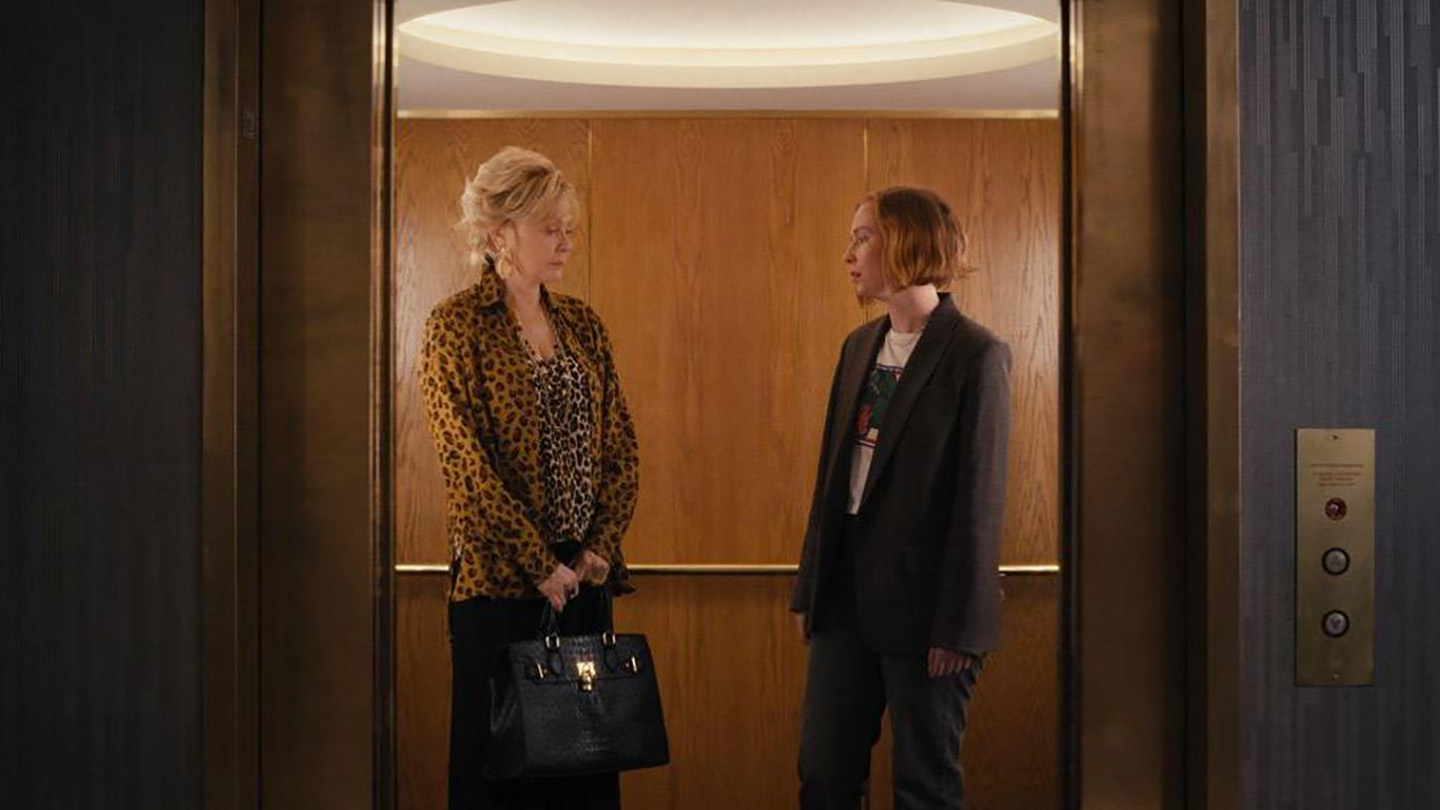 Two characters from a film standing in an elevator, one in a leopard print coat and the other in a blazer