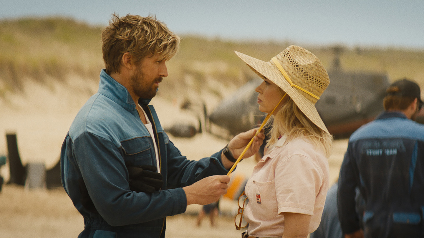 Two actors on set, man in denim jacket and woman in straw hat, in a beach scene, having a conversation