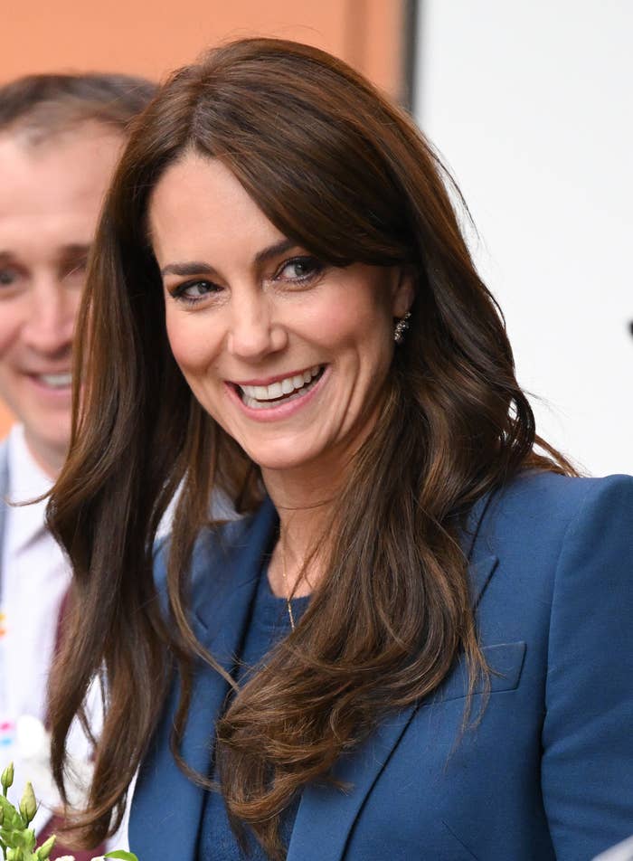 Kate Middleton smiling, wearing a blazer with long wavy hair, at a public event