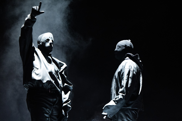 Two masked performers on stage, one with a raised hand gesture, in dark outfits against a smoke-filled backdrop