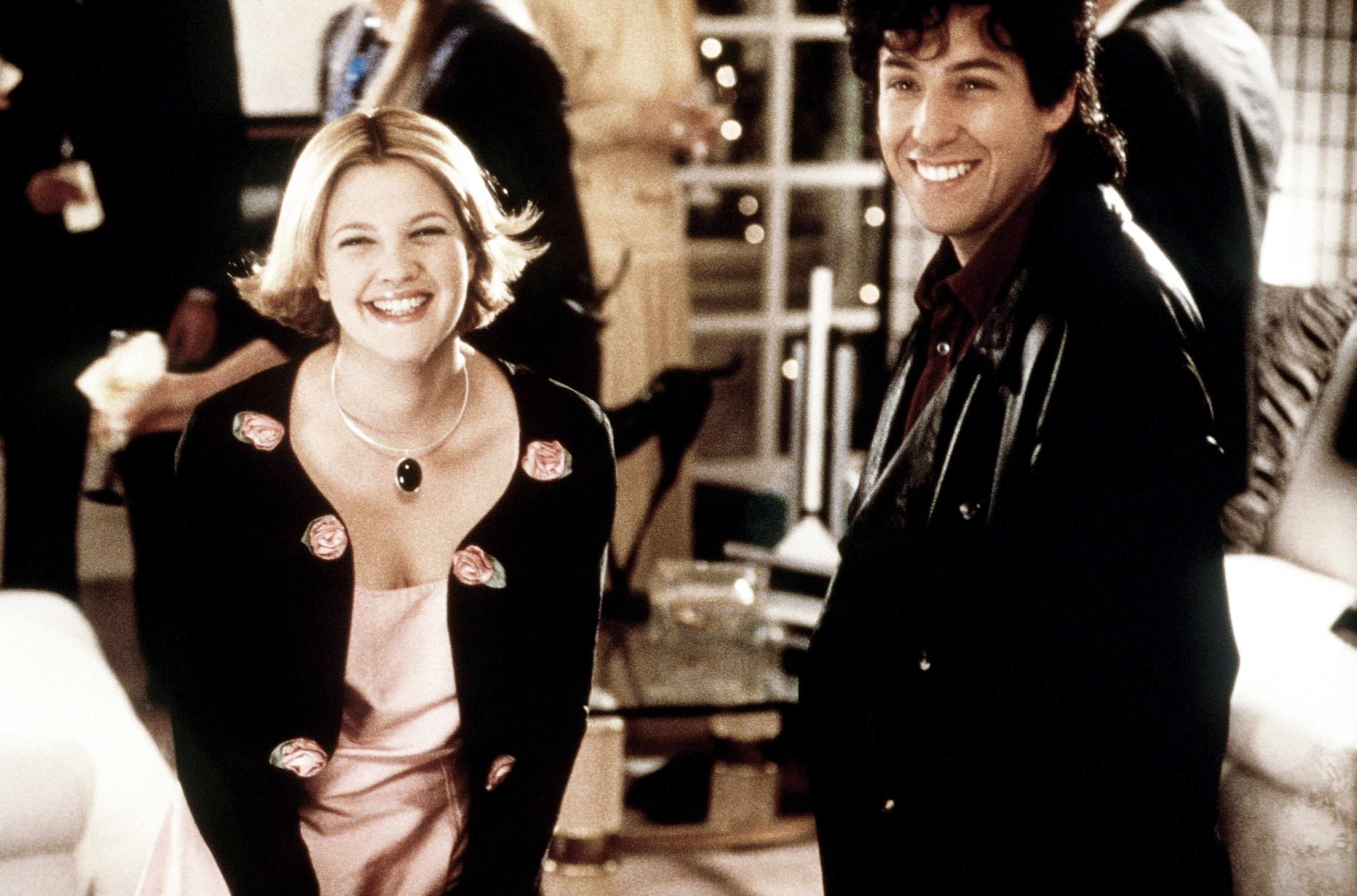 Drew Barrymore in a pink dress and Adam Sandler in a leather jacket smiling in a scene from a rom-com movie