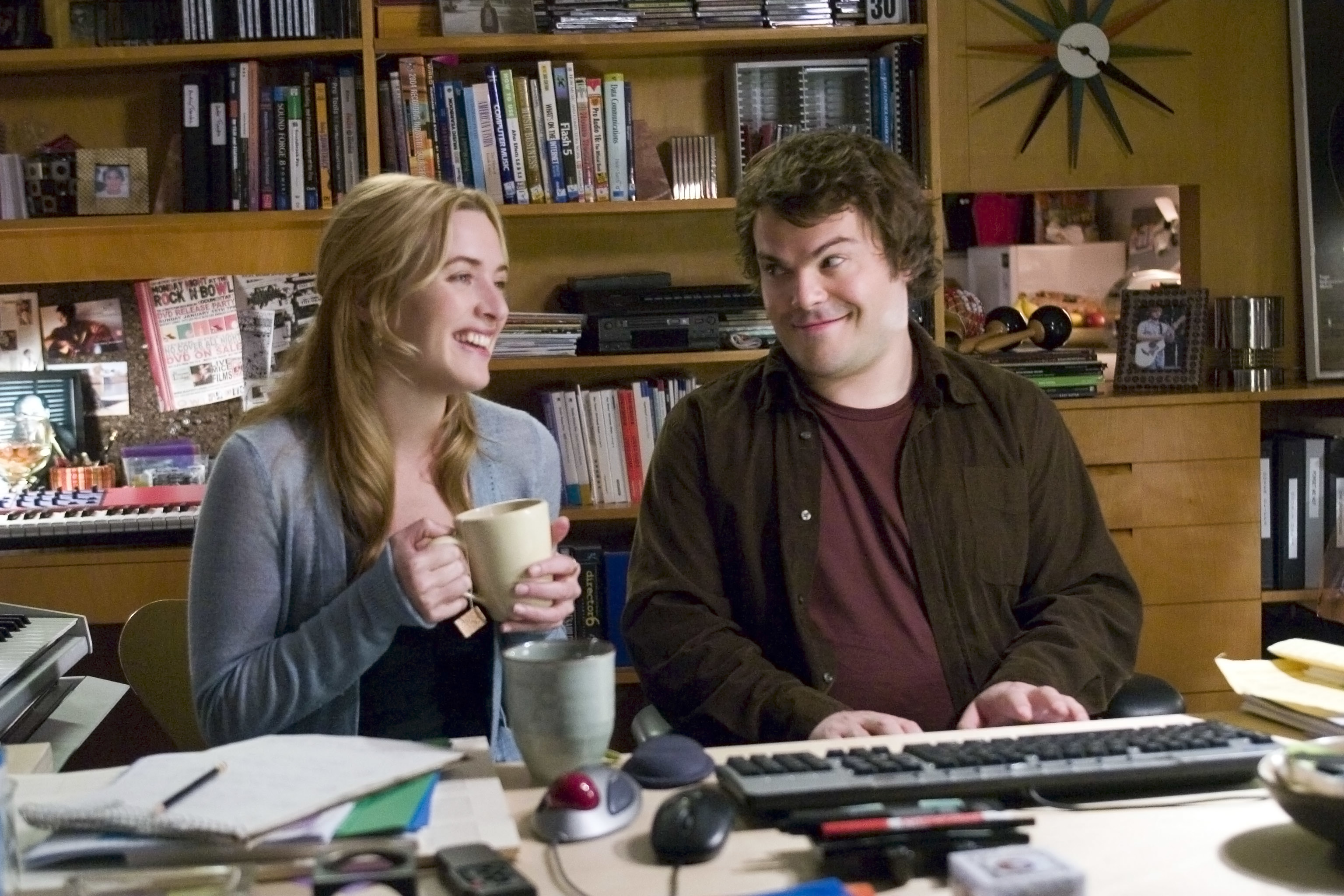 Two characters from a film in an office setting, sharing a light moment by a computer desk