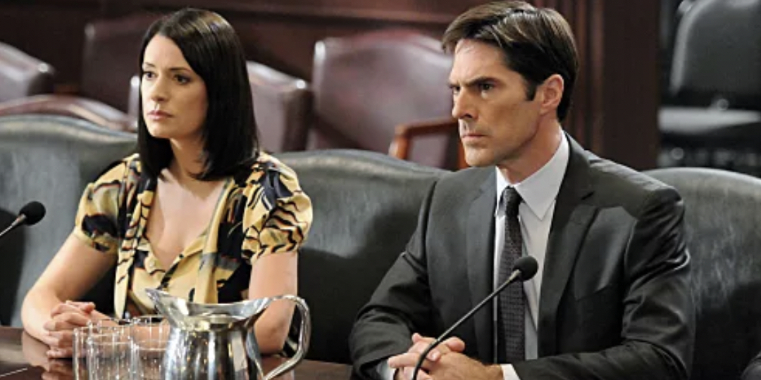 Two actors in a courtroom setting on a TV show, man in a suit and woman in a patterned top, both seated