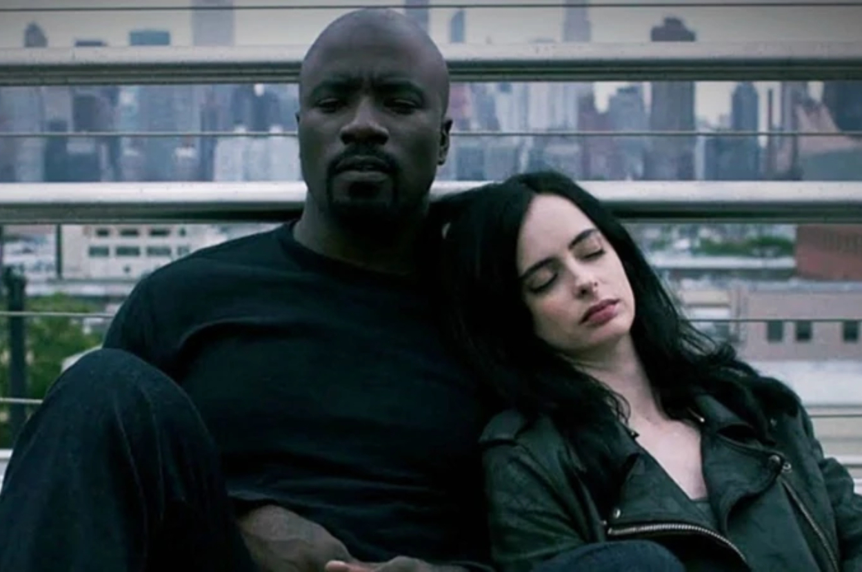 Characters Luke Cage and Jessica Jones sitting close, with a city background, in a scene from their TV show