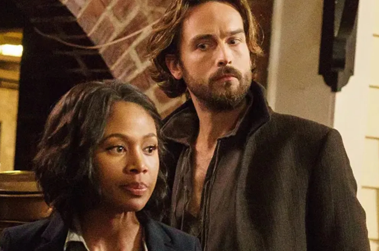 Two actors in a scene from a TV show, man with beard and woman in a jacket, looking serious