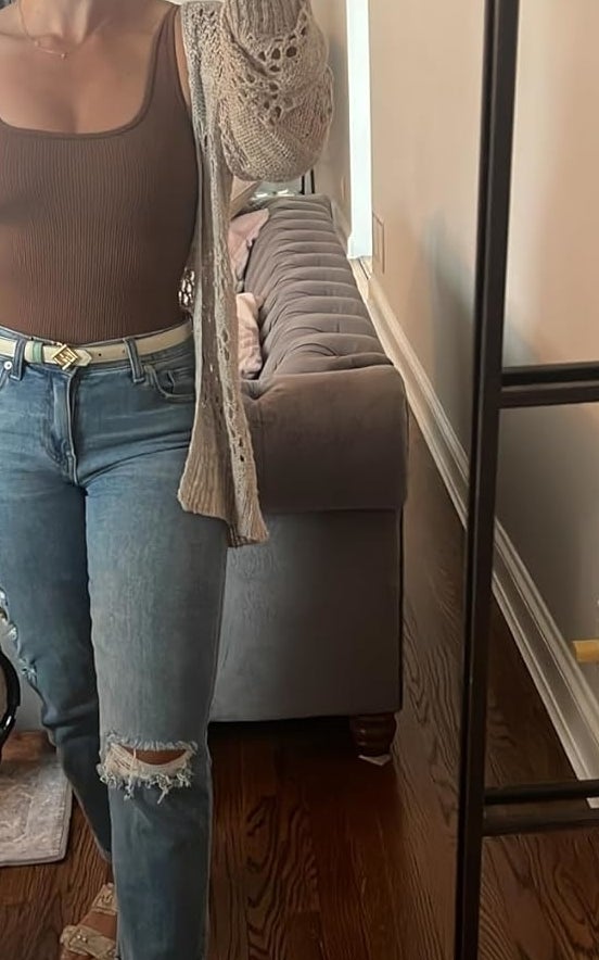 person in a mirror selfie wearing a brown top, distressed jeans, a belt, and heeled sandals, holding a phone and a bag