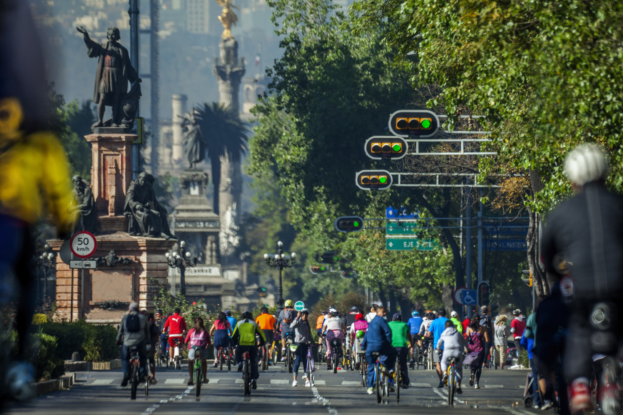 Cyclists ride on a city street with traffic lights and a statue in the background