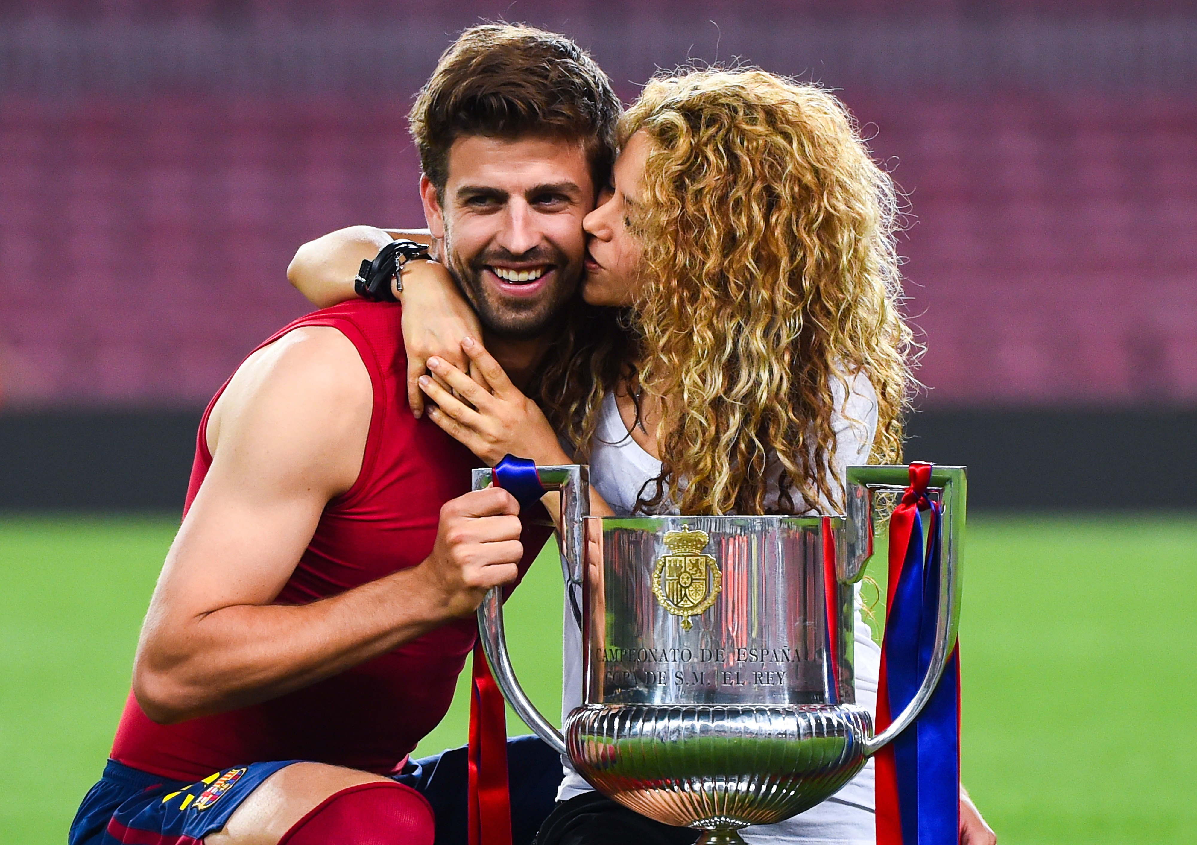 Shakira kisses Gerard Piqué who smiles, holding a trophy on a sports field